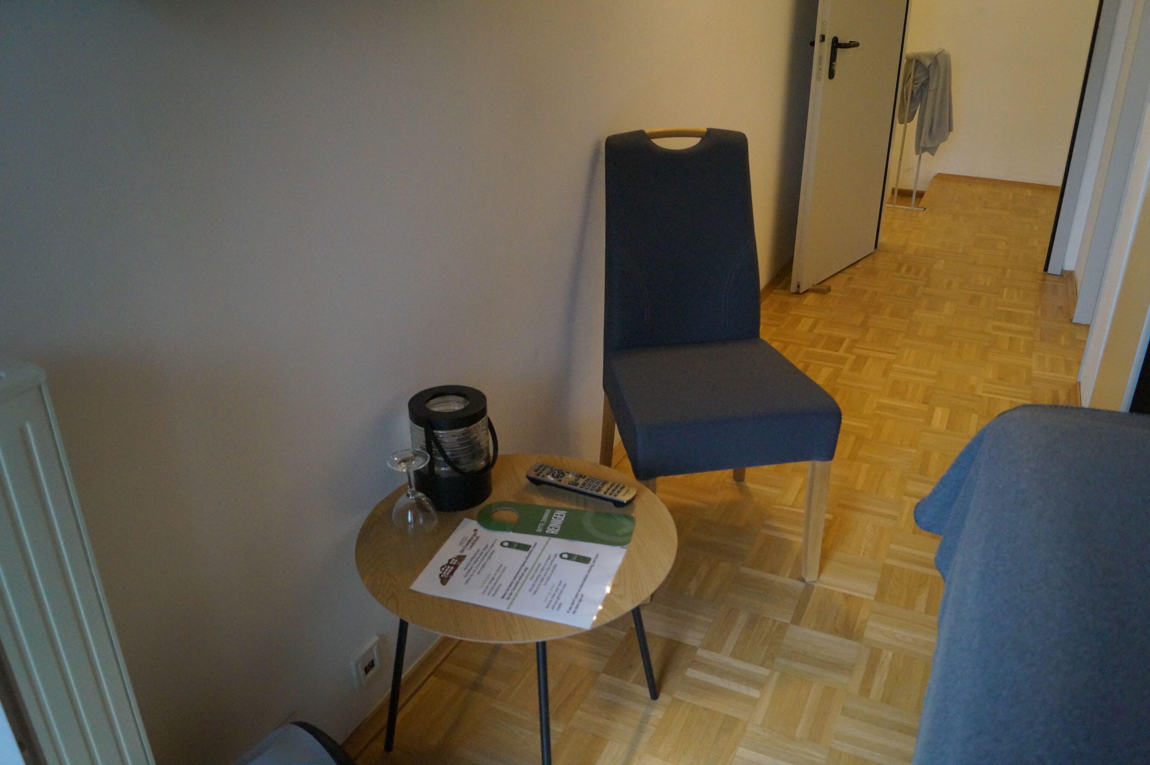 A small room with a wooden floor, a round table with a glass, a remote control, and a chair.