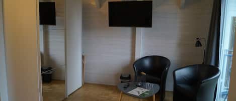 Una habitación con suelo de parqué en espiga, un televisor de pantalla plana montado en la pared, dos sillas negras, una mesita redonda con un vaso y un mueble de espejo.
