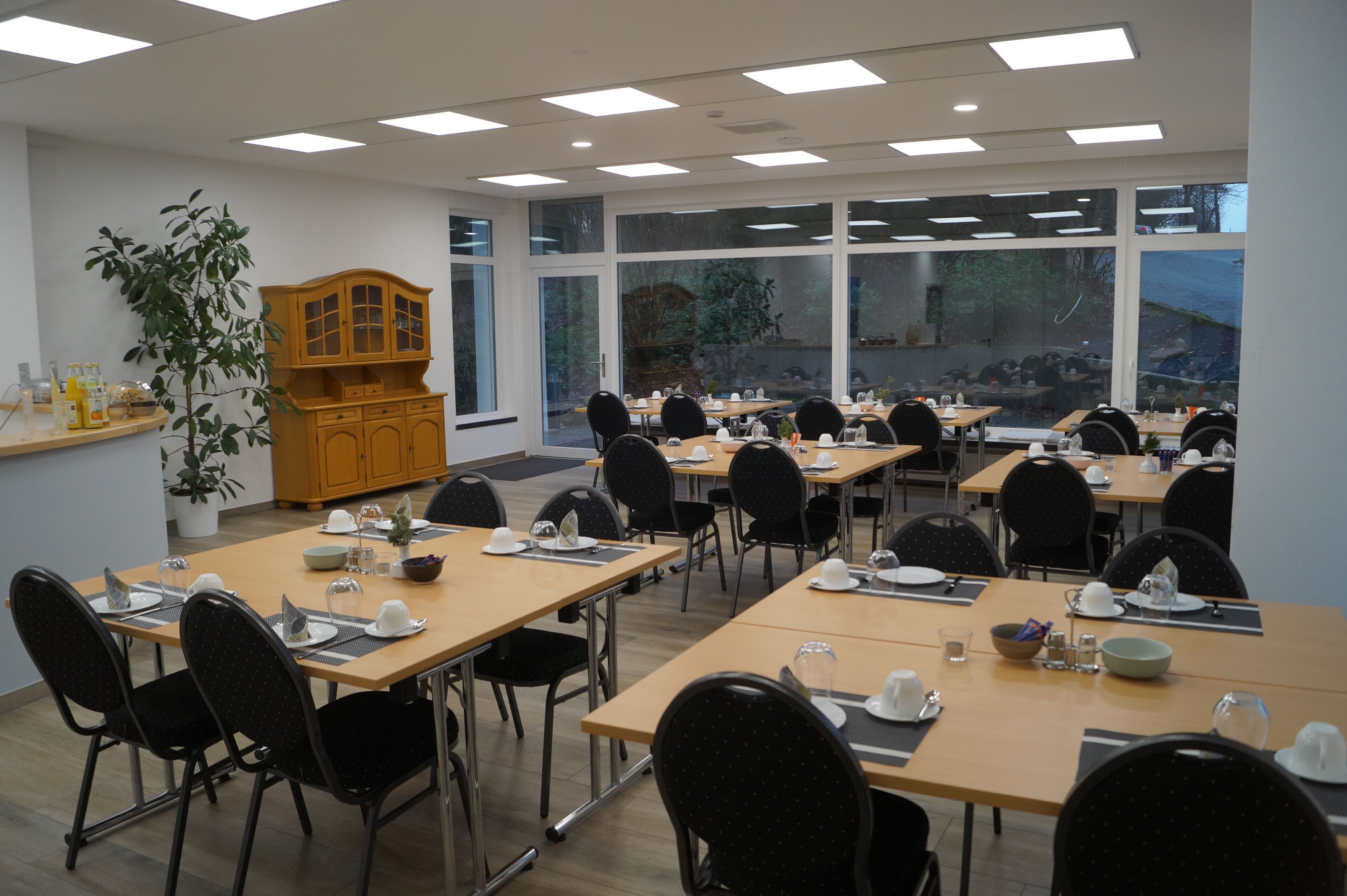 A conference room with multiple tables and chairs, large windows, and a wooden cabinet with a plant.