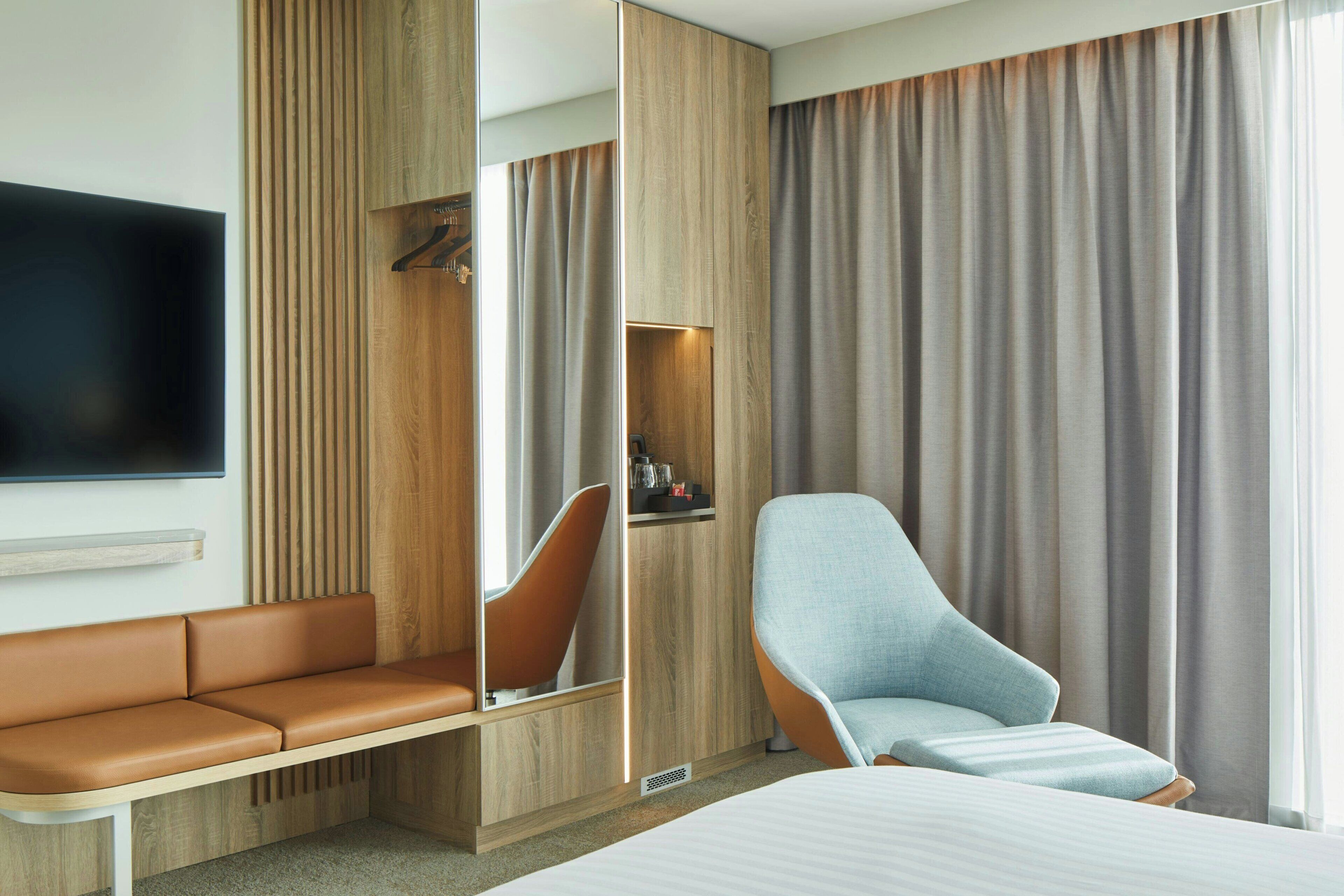 Una habitación de hotel moderna con una cama grande, una zona de estar con sofá y sillones, un televisor montado en la pared y un armario con espejo.
