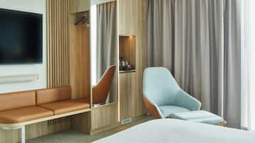 A modern hotel room with a large bed, a seating area with a sofa and armchairs, a TV mounted on the wall, and a wardrobe with a mirror.
