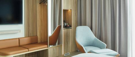 Una habitación de hotel moderna con una cama grande, una zona de estar con sofá y sillones, un televisor montado en la pared y un armario con espejo.