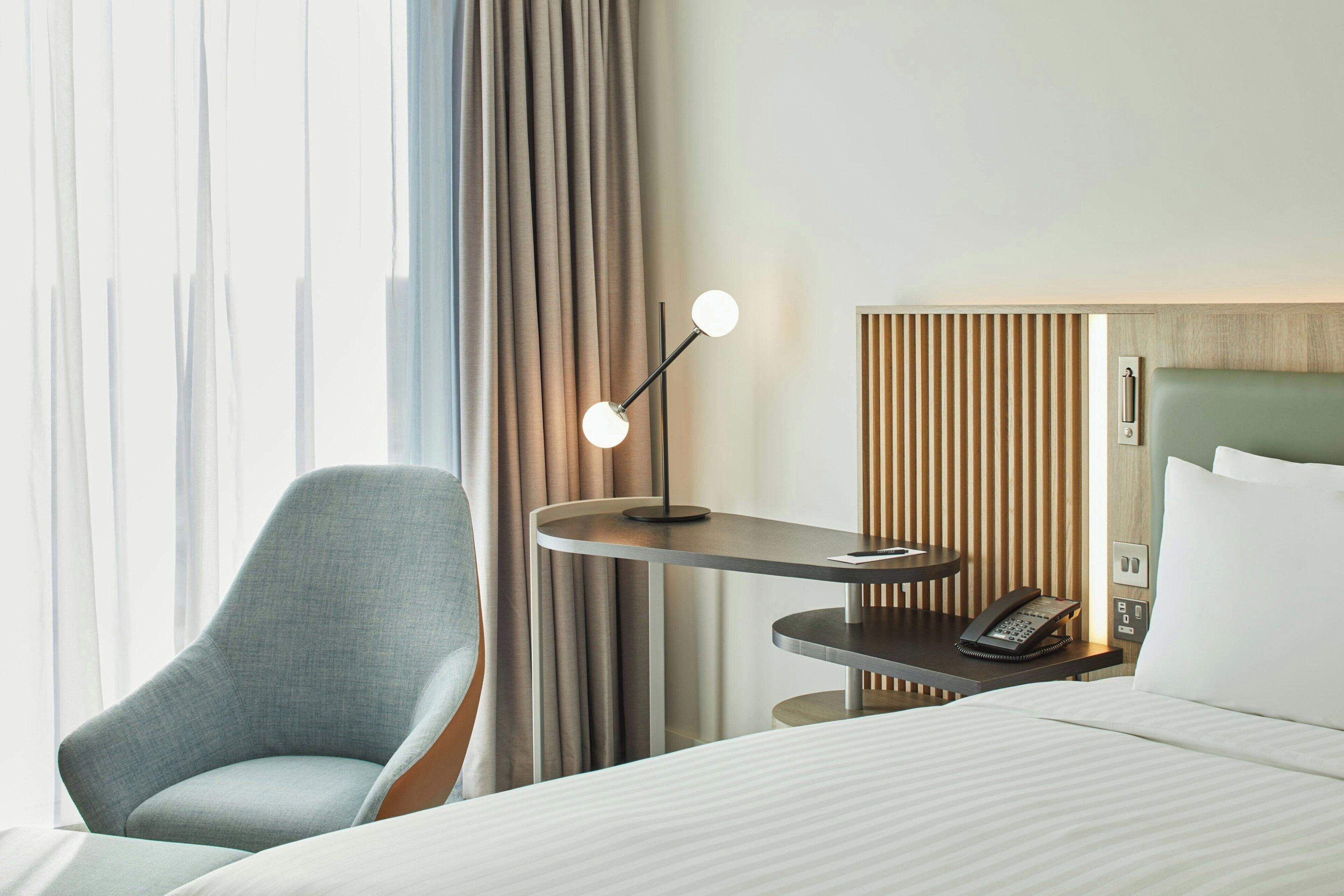 A hotel room with a bed, a desk with a phone, a chair, and a lamp.