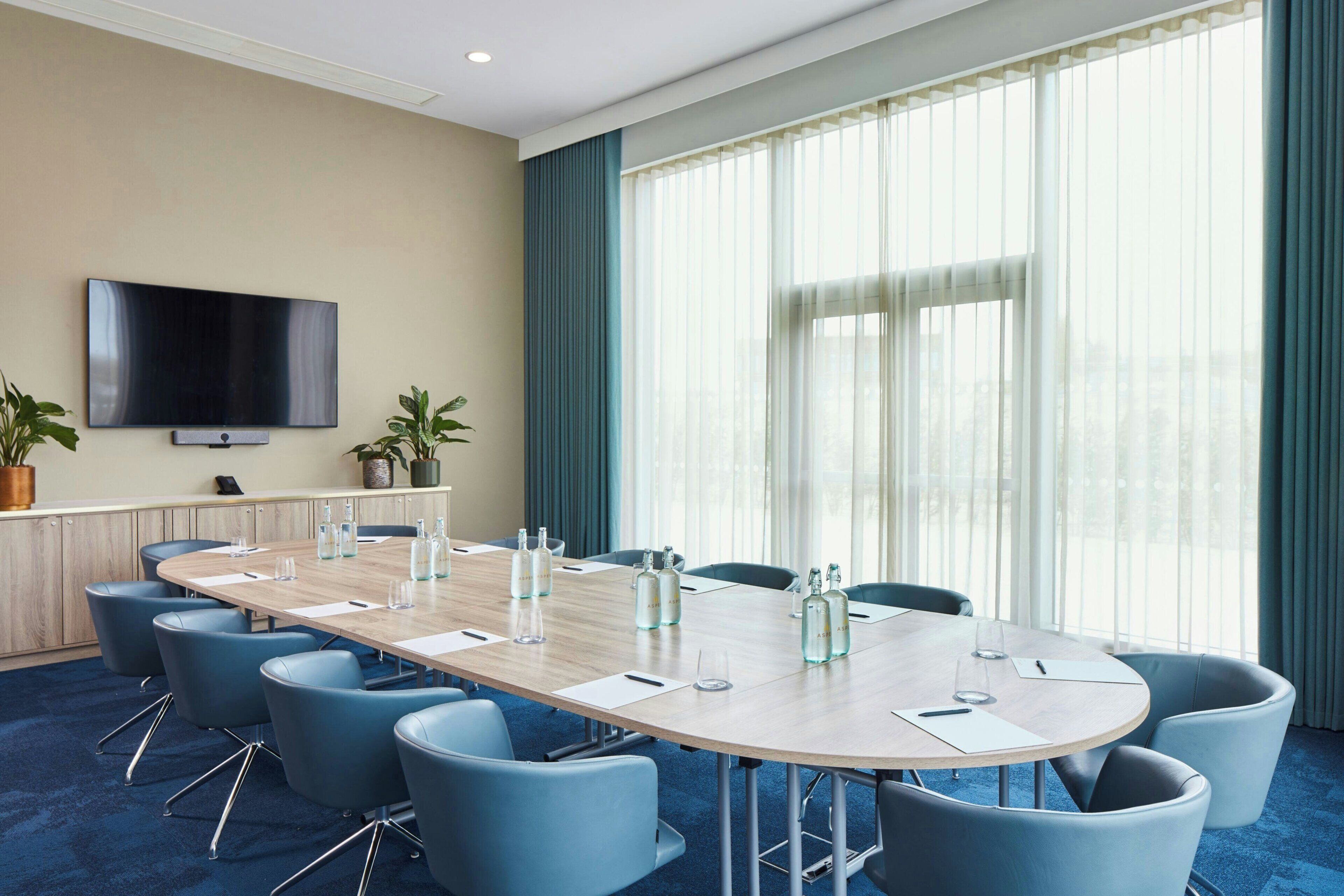 A conference room with a long table, blue chairs, a flat-screen TV, and large windows with curtains.