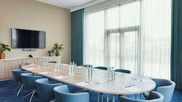 A conference room with a long table, blue chairs, a flat-screen TV, and large windows with curtains.