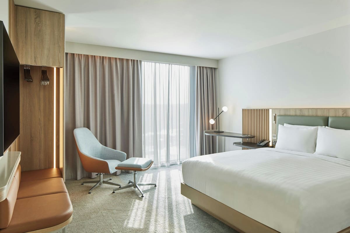 Quarto de hotel com uma cama grande, área de estar com uma poltrona e apoio para os pés, escrivaninha com luminária e televisão na parede.