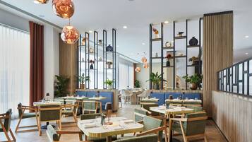 Een modern restaurantinterieur met ronde tafels, beklede stoelen, hanglampen en diverse decoratieve objecten op planken.