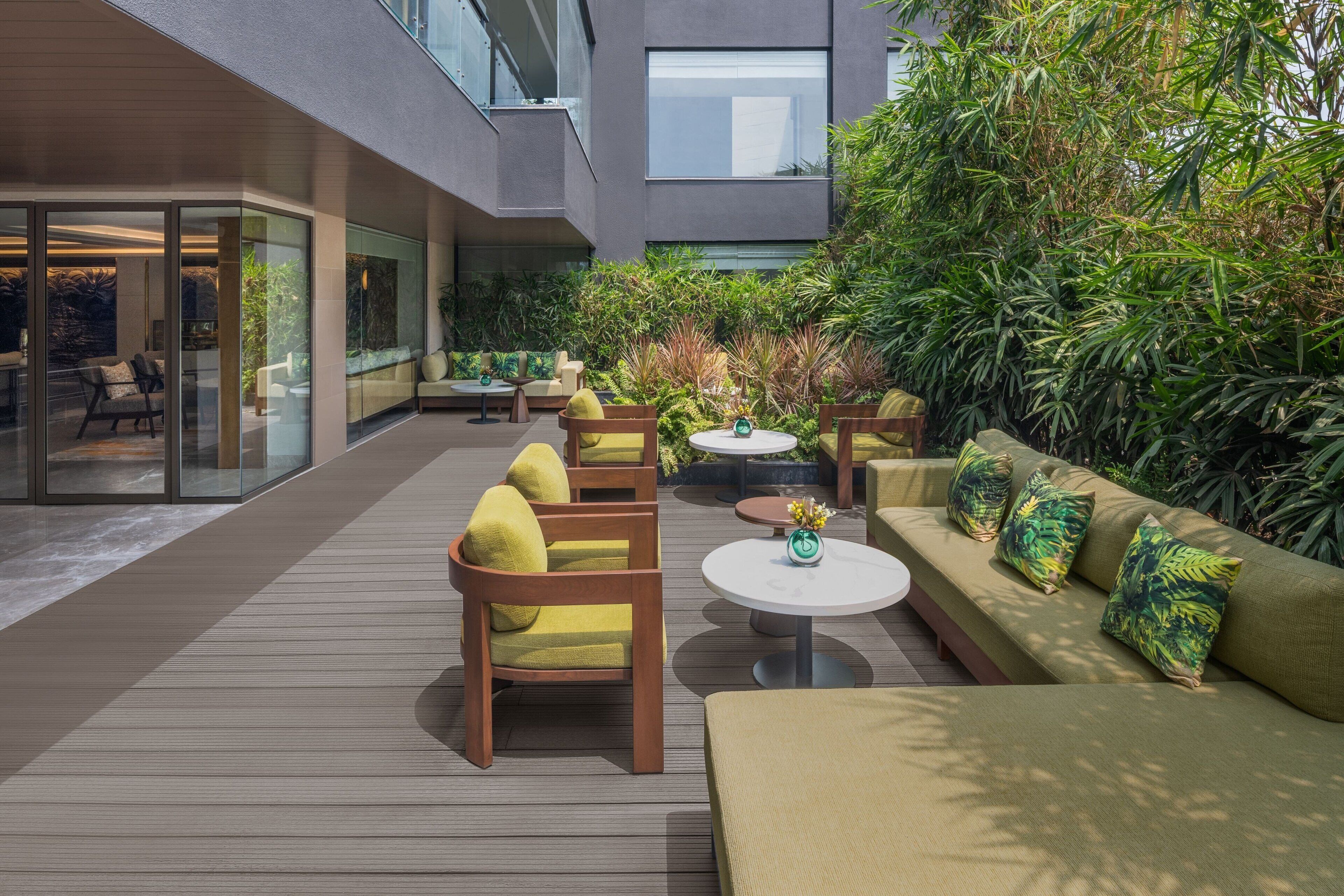 A modern outdoor patio with wooden furniture, a green sofa, and a glass wall overlooking the interior.