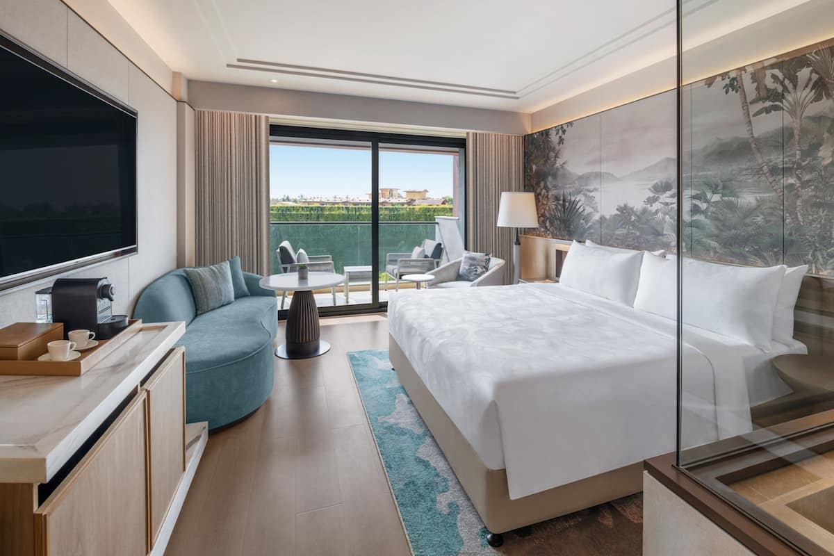 A modern hotel room with a large bed, a sofa, a coffee station, a balcony with a view, and a large TV.