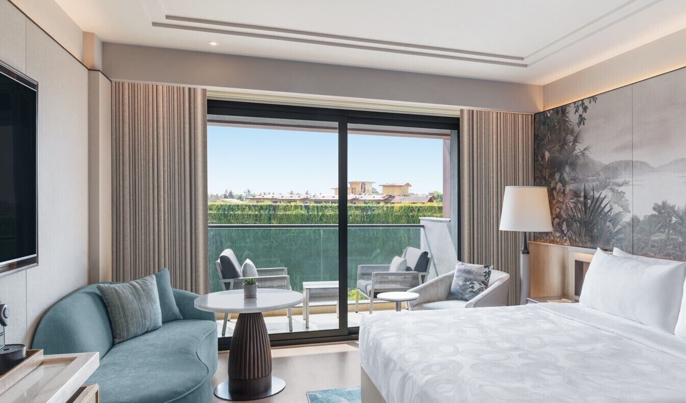 A hotel room with a large bed, a sofa, a small table, and a balcony with a view of the city.