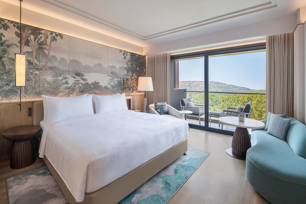 A modern hotel room with a large bed, a sofa, a dining table, and a balcony with a view of the landscape.
