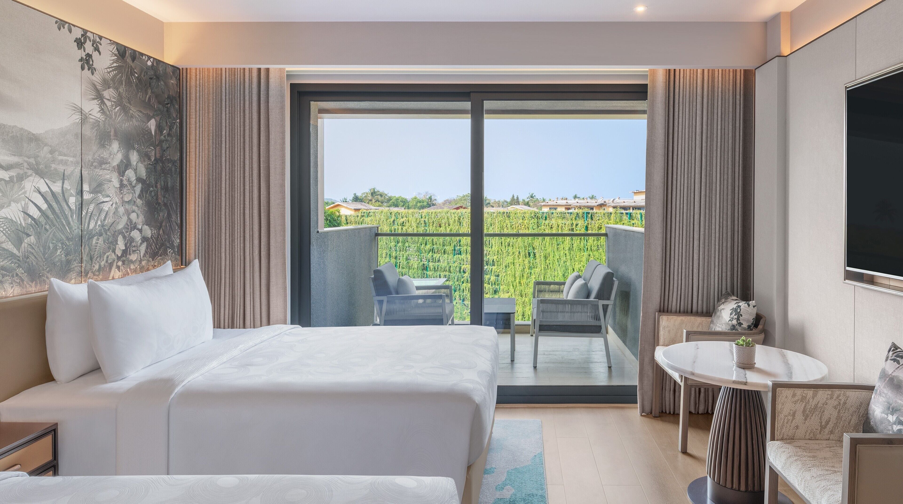 A hotel room with a bed, a small table, a chair, and a balcony with a view of greenery and buildings.