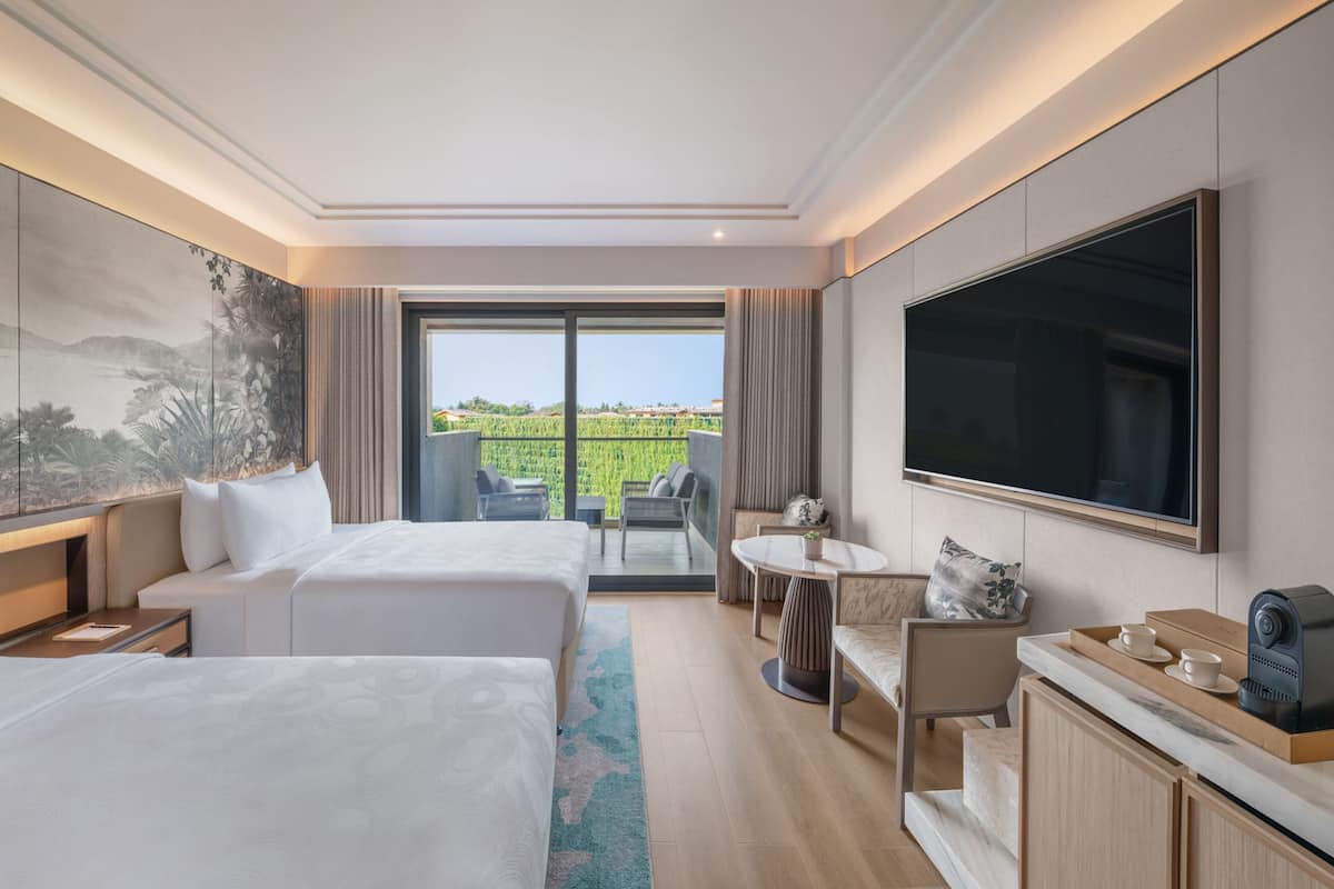 A hotel room with two beds, a large TV, a dining area with a table and chairs, and a view of a green landscape through the window.