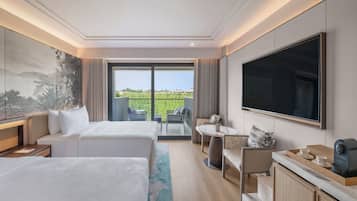 A hotel room with two beds, a large TV, a dining area with a table and chairs, and a view of a green landscape through the window.