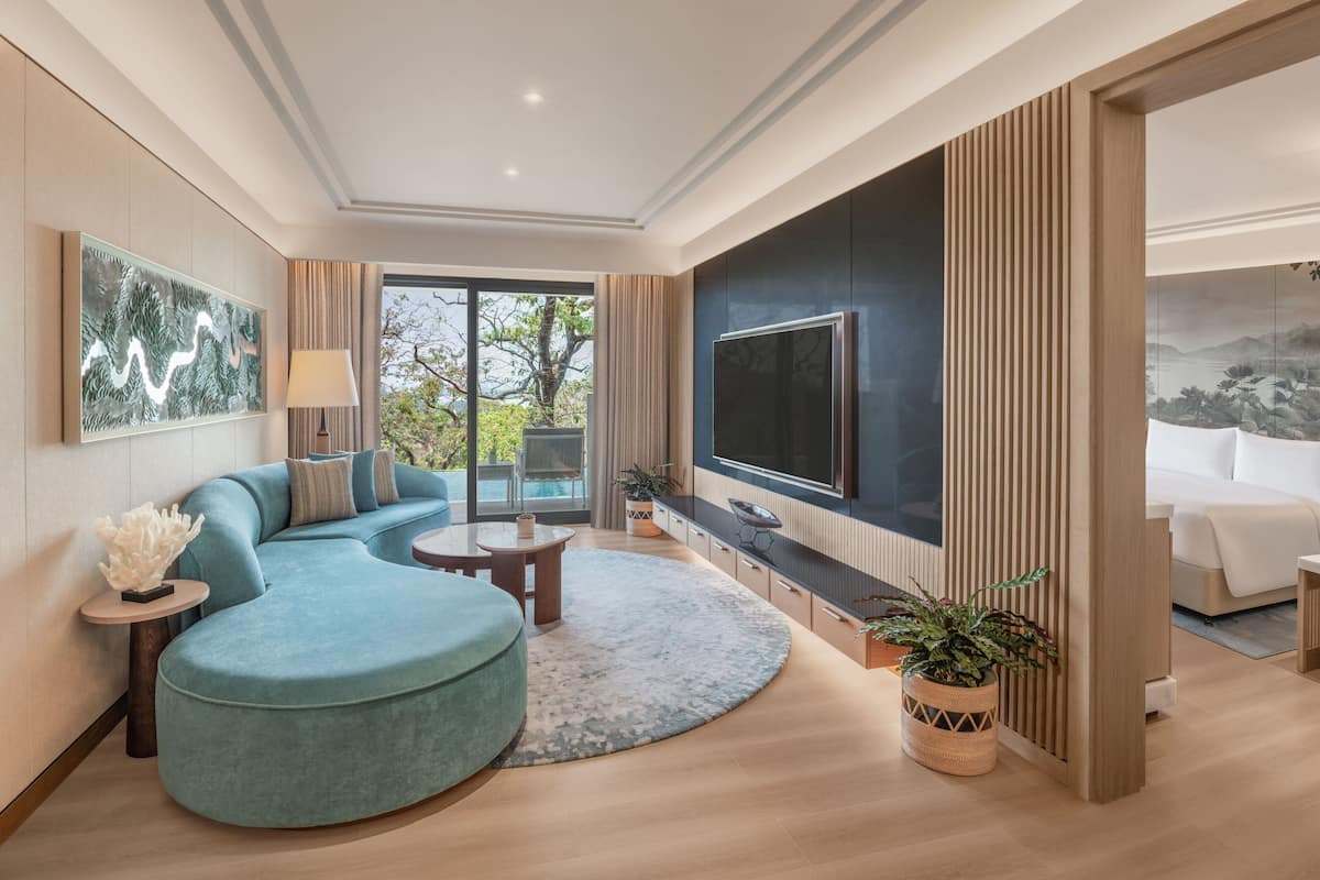 A modern living room with a curved sofa, a flat-screen TV, and a large window offering a view of greenery.