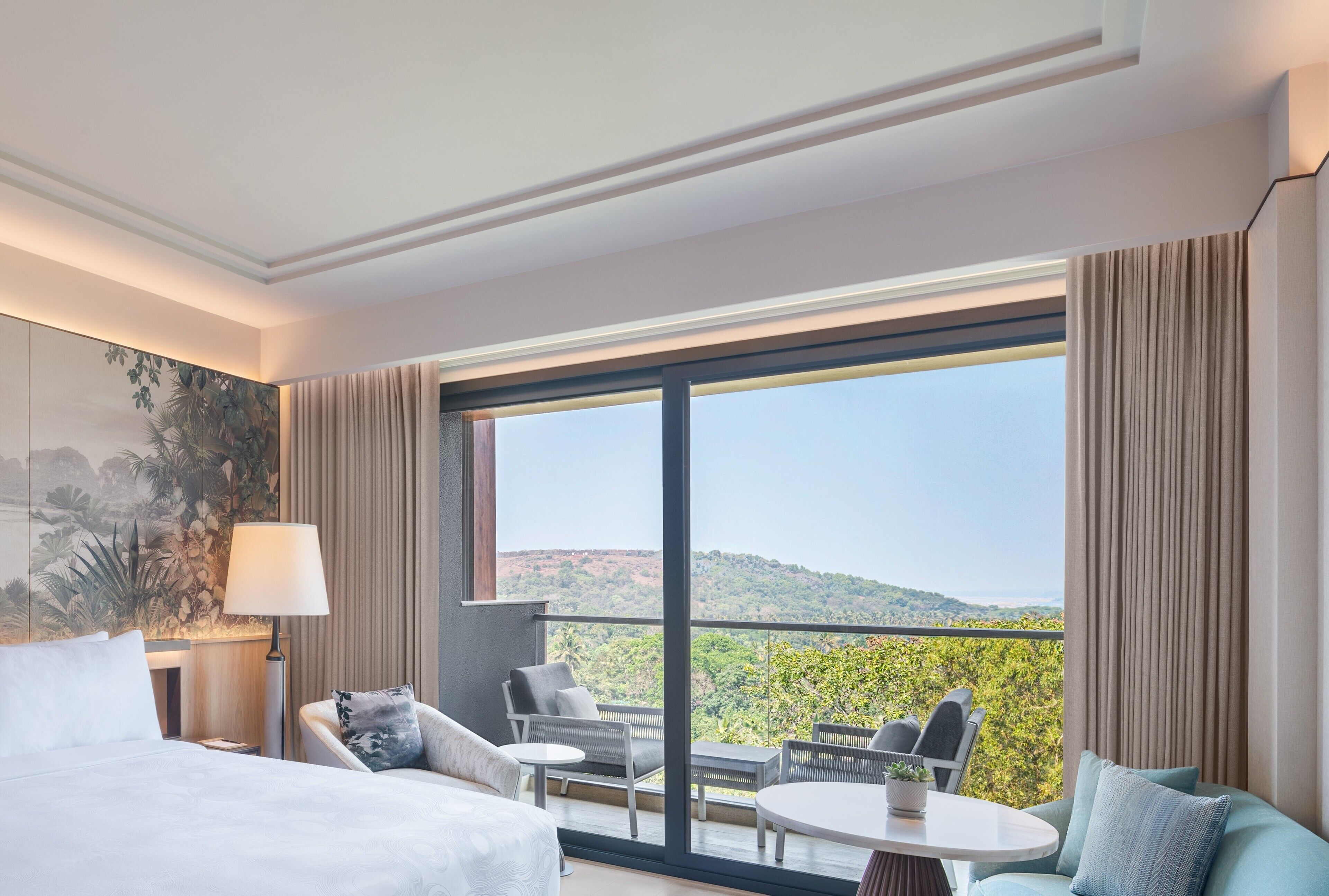 A hotel room with a large window, a bed, a sofa, a table, and a lamp.
