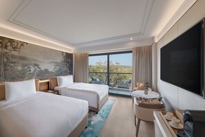A hotel room with two beds, a desk with a chair, a TV, and a balcony with a view of the landscape. - JW Marriott Goa (Vagator)
