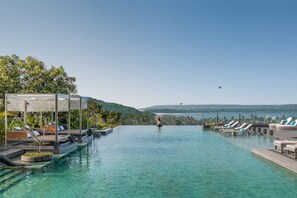 Un espace piscine doté de chaises longues et d’un auvent, entouré de verdure et avec des collines en arrière-plan.