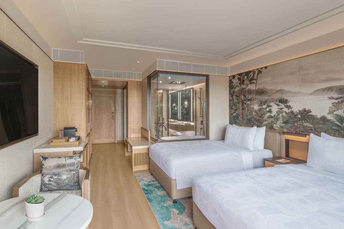 A modern hotel room with two beds, a sitting area, a bathroom with a glass shower, and a large mural on the wall.