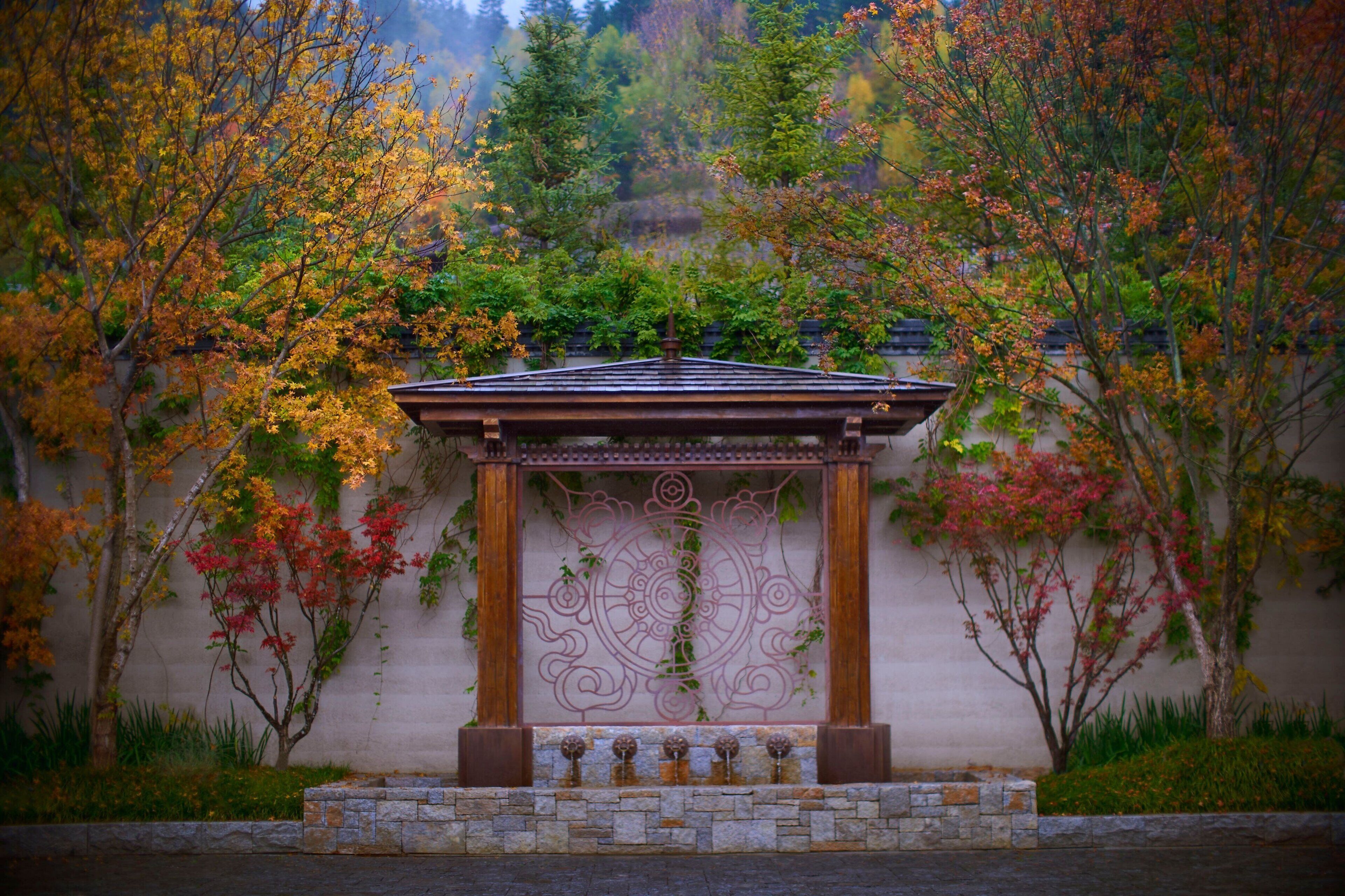 A gazebo with a stone base and a metal railing, surrounded by trees with autumn foliage.