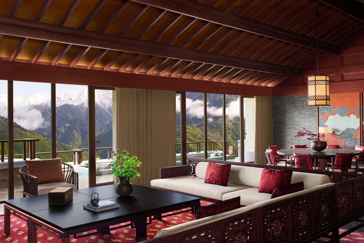 Amplia habitación con un ventanal que ofrece vistas a la montaña, un sofá con cojines rojos, una mesa de centro de madera y un comedor con sillas y mesa rojas.
