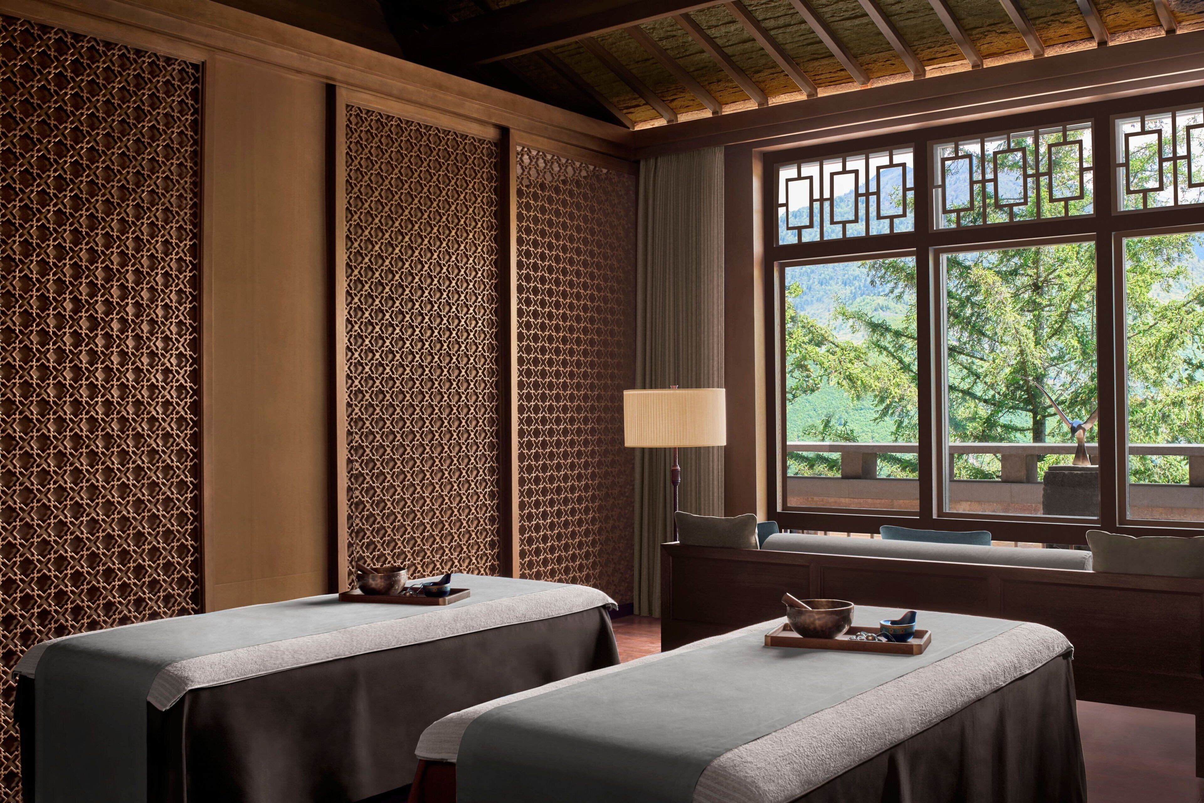 Ein Raum mit zwei Massagetischen, einem großen Fenster mit Blick auf Bäume und einer Wand mit traditionellem Muster.