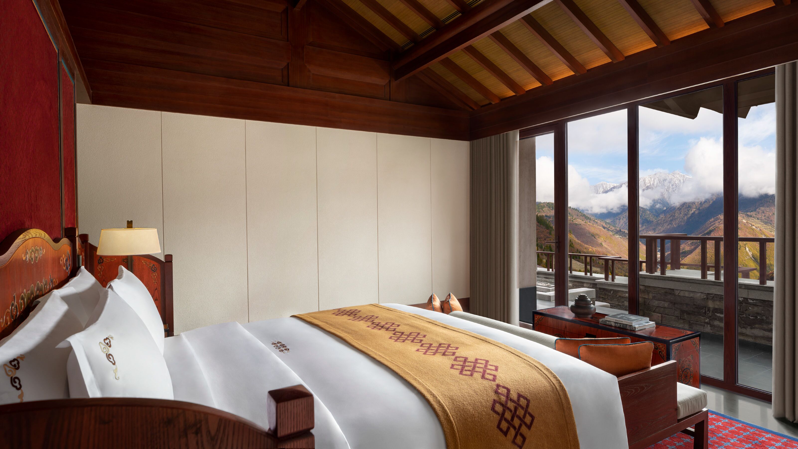 A hotel room with a bed, a nightstand, a lamp, and a view of mountains through large windows.