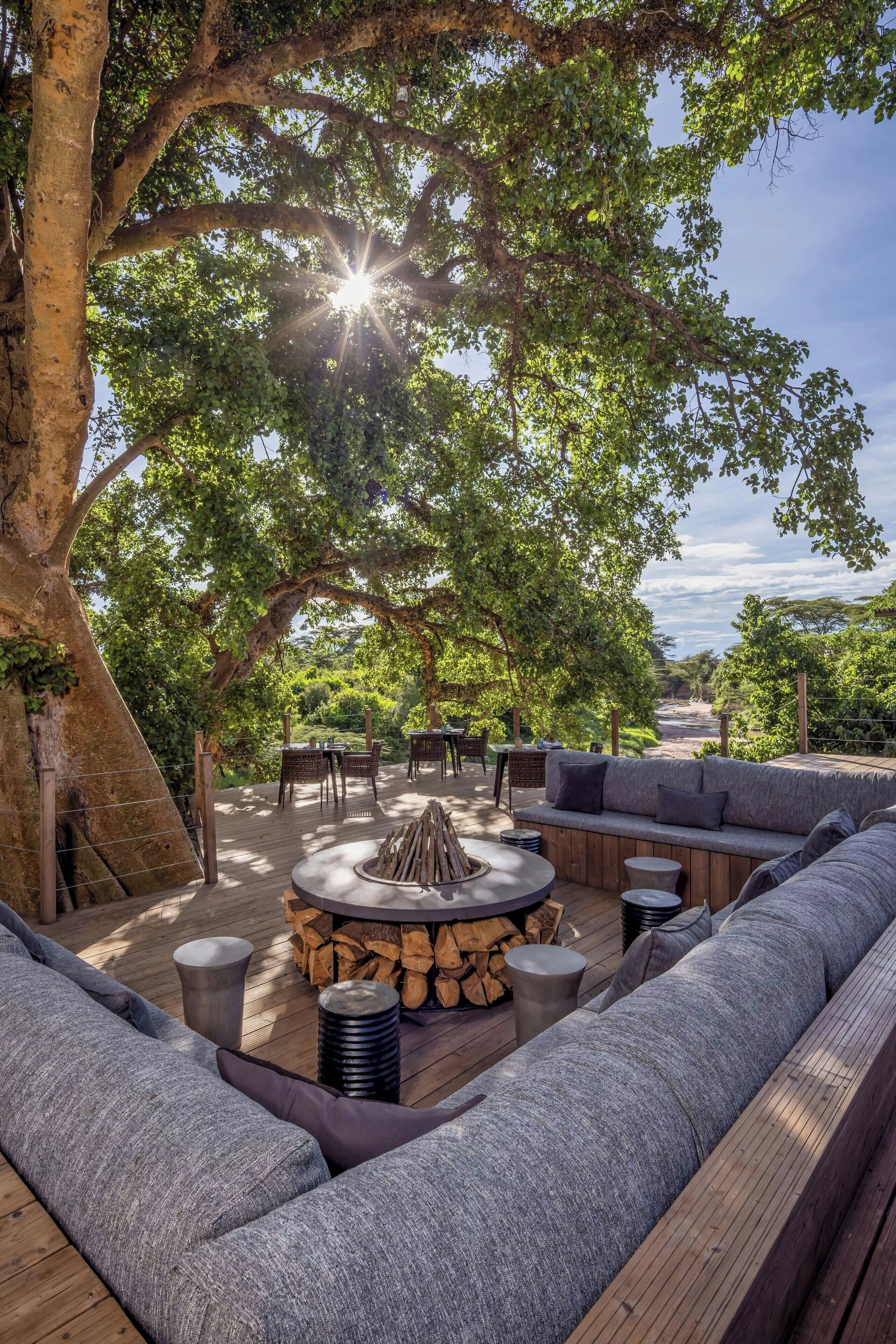 An outdoor seating area with a fire pit, wooden deck, and a large tree providing shade.