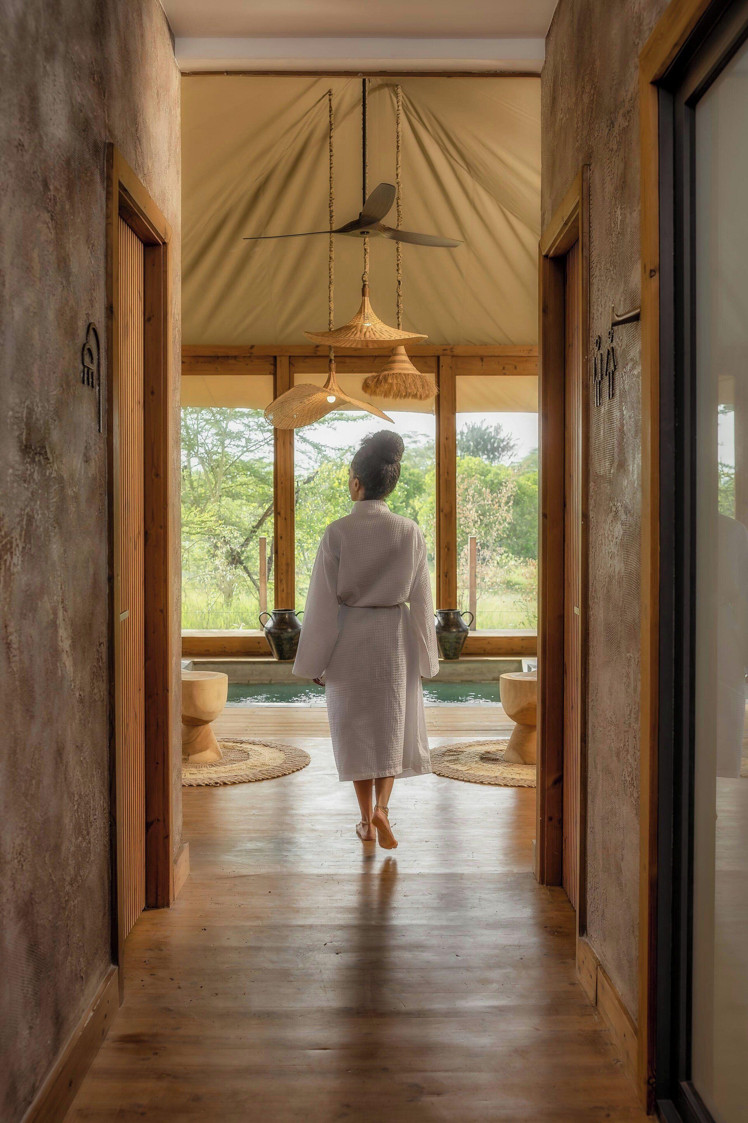 A person in a white robe walking down a wooden hallway towards a window with a view of greenery.