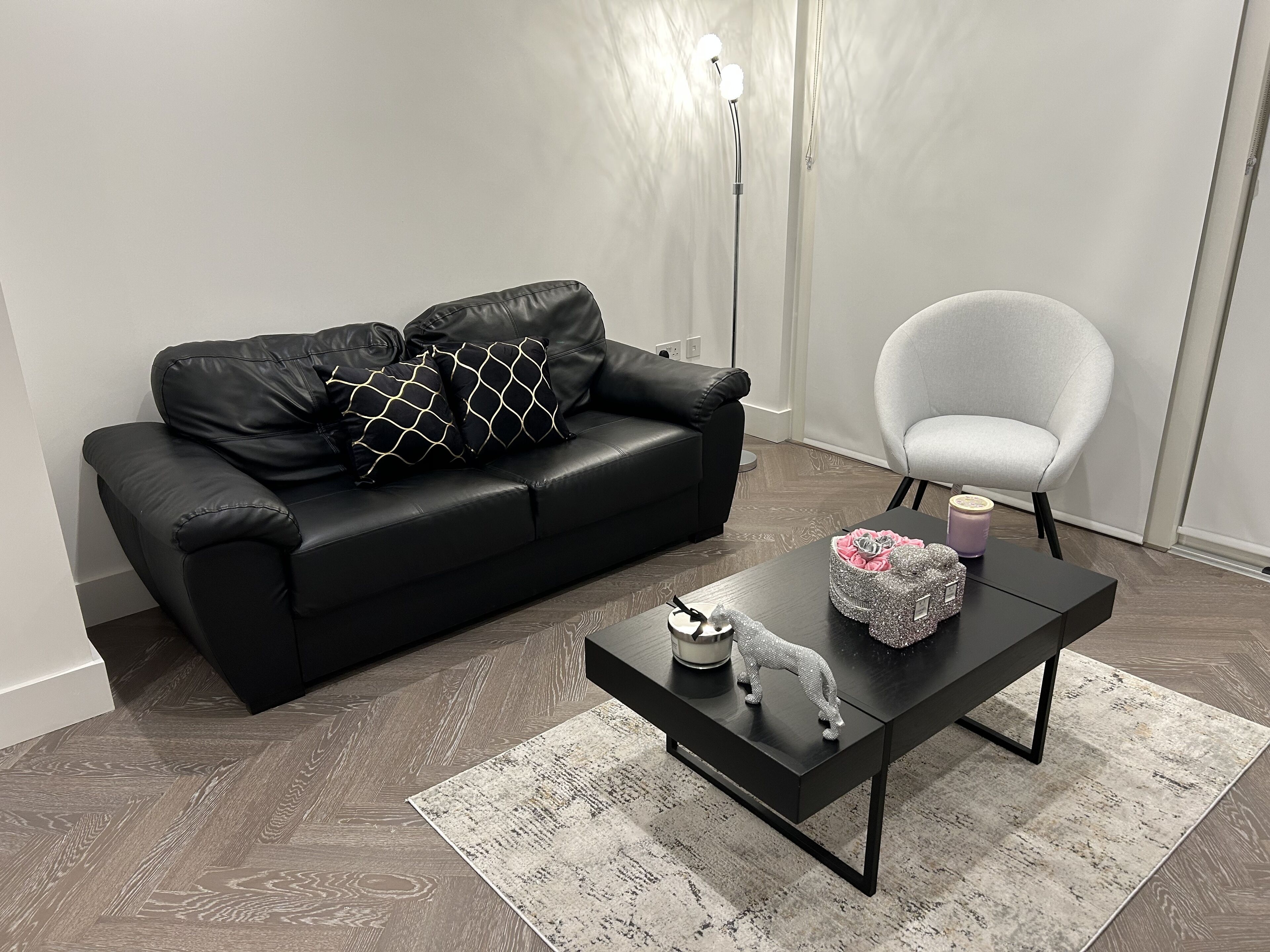 Una sala de estar moderna con un sofá de cuero negro, un sillón blanco, una mesa de centro negra con objetos decorativos y una alfombra estampada.