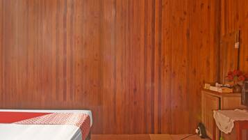 A room with a bed, a wooden wall, a small table with a vase of flowers, and a mirror.