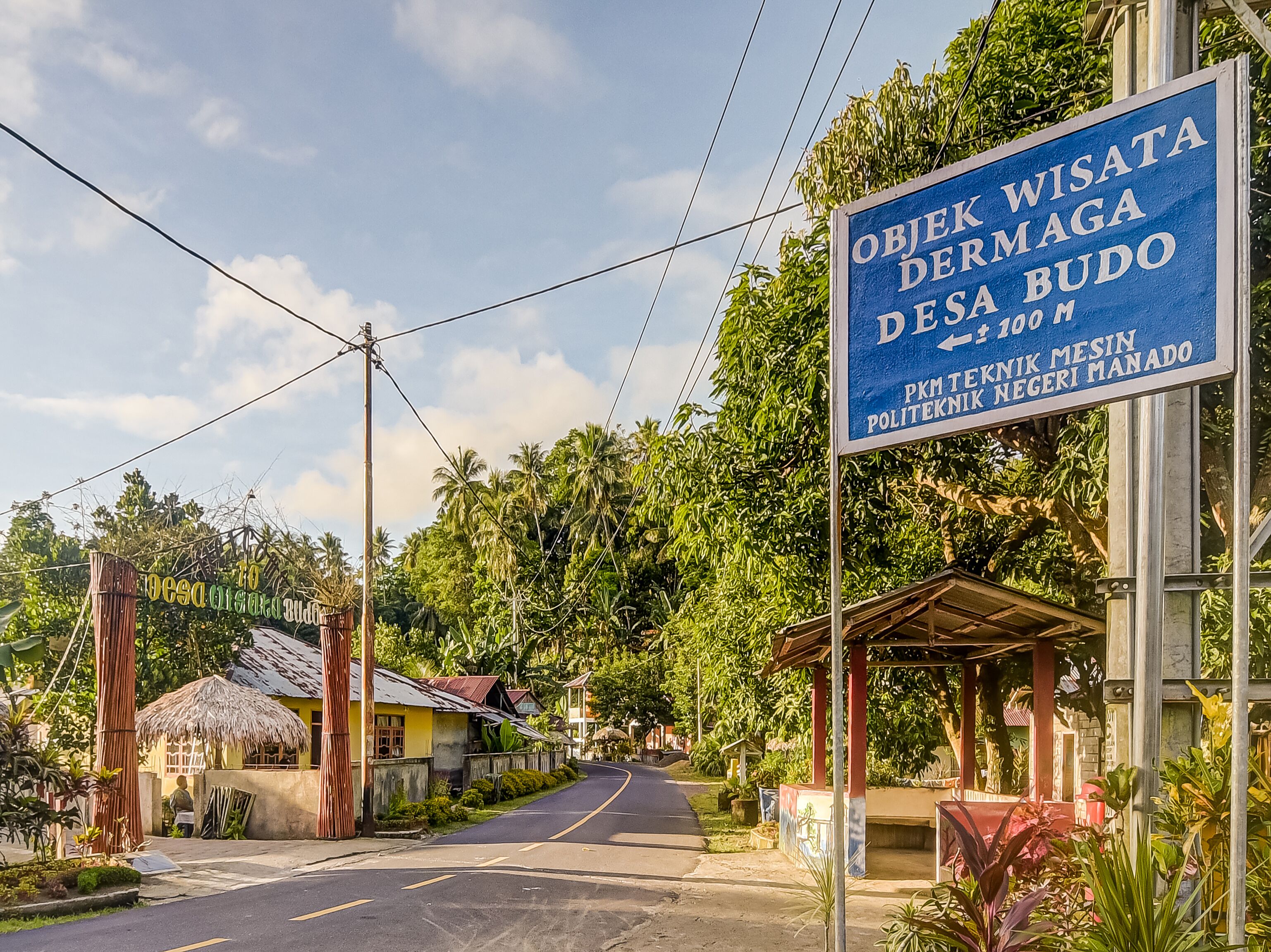 Une vue de rue avec une enseigne indiquant un hôtel nommé « Objek Wisata Dermaga Desa Budo » et un petit bâtiment doté d’un toit de chaume.