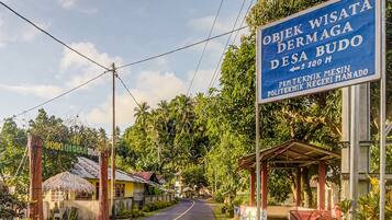Une vue de rue avec une enseigne indiquant un hôtel nommé « Objek Wisata Dermaga Desa Budo » et un petit bâtiment doté d’un toit de chaume.
