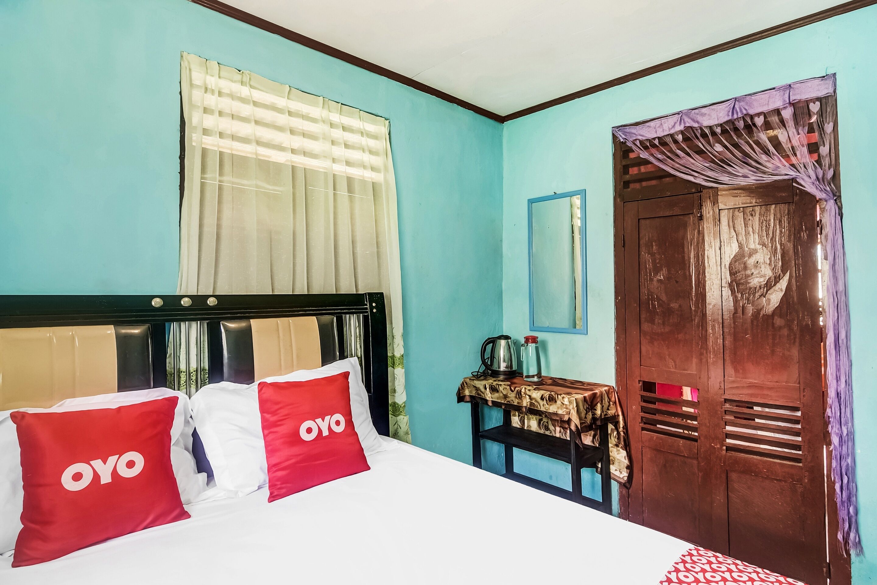 A hotel room with a bed, a wooden wardrobe, a small table with a kettle, and a window with curtains.