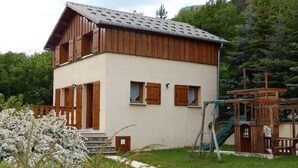 A two-story building with a wooden exterior and a white facade, surrounded by greenery and a playground. - 6-person chalet in the heart of Champsaur (ST JEAN ST NICOLAS)