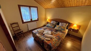 A bedroom with a wooden ceiling, a bed with floral bedding, two bedside lamps, a chair, and a window with curtains. - 6-person chalet in the heart of Champsaur (ST JEAN ST NICOLAS)