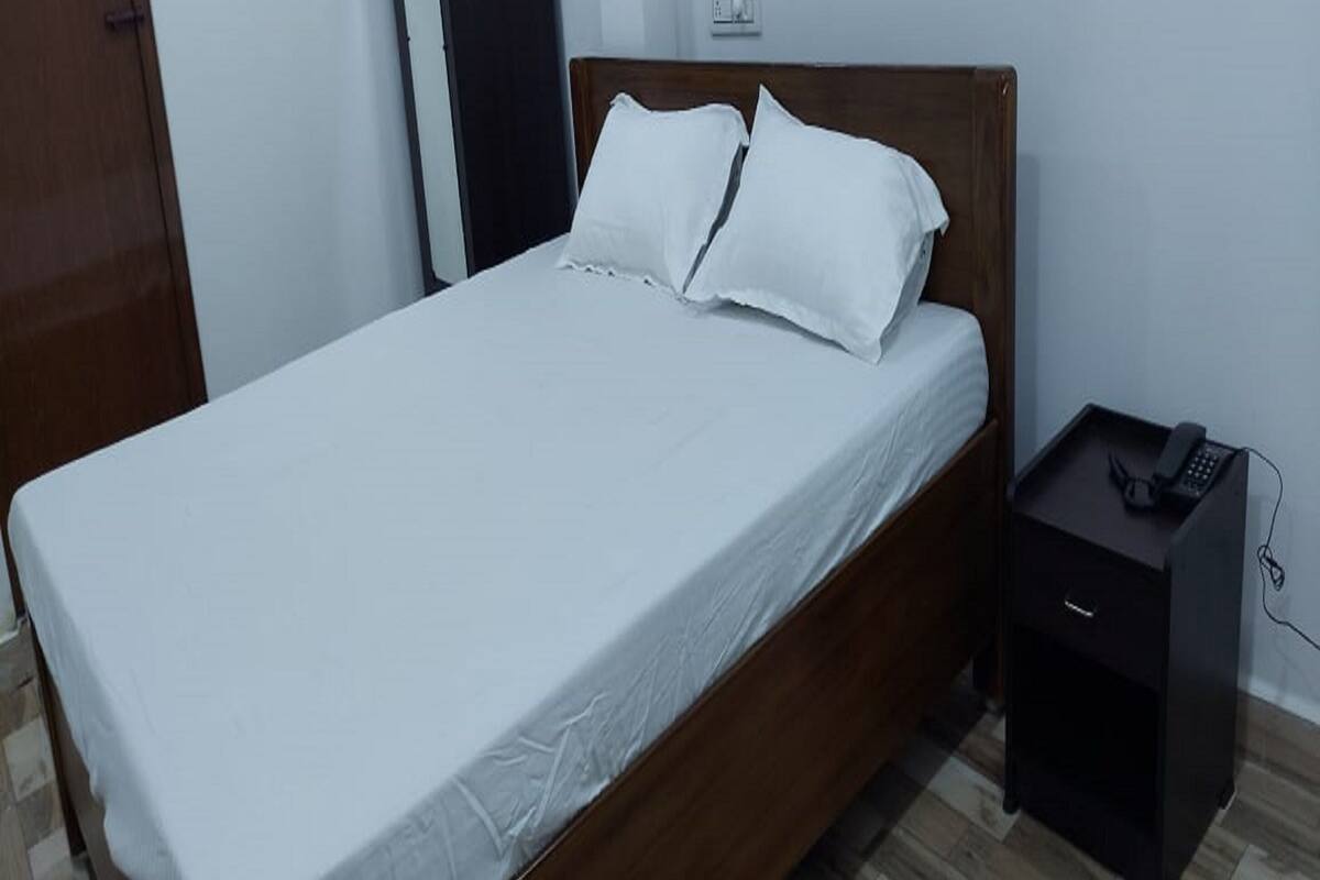 Un lit simple avec un cadre en bois et du linge de lit blanc, une petite table de chevet avec un téléphone et une prise électrique murale.