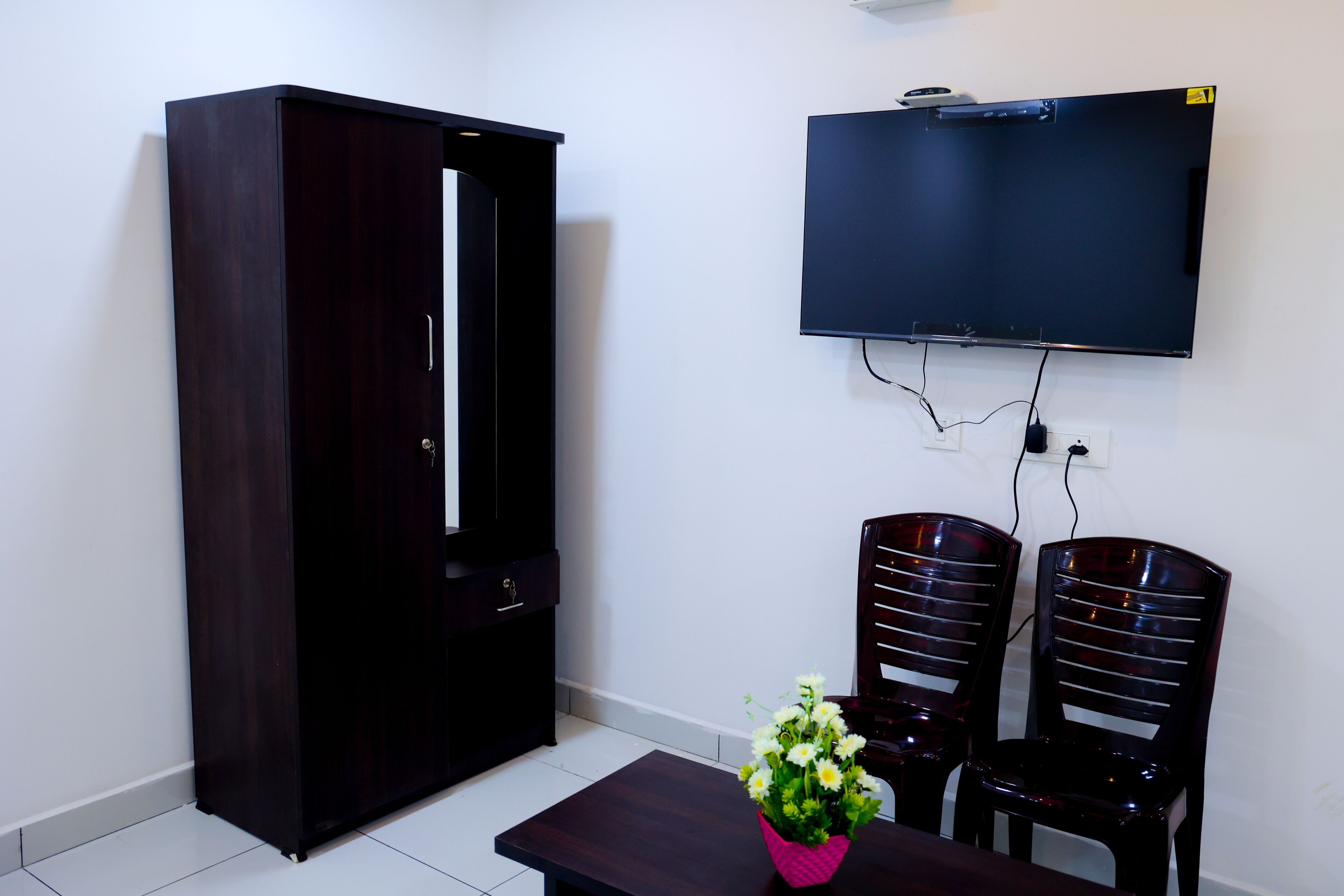 A room with a dark wooden wardrobe, a flat-screen TV mounted on the wall, two black chairs, and a small table with a pink vase of flowers.