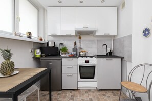 A compact kitchen with white cabinets, a stove, a sink, and a small dining area with a table and chairs. - Stylish Apartment with Balcony | Close to Beach and Park (Gdansk)