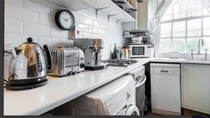 A kitchen with white appliances, a clock, and a window with a view of greenery. - Charming Studio Sleeps 2 - Kentish Town Nwlondon (London)