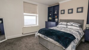 A bedroom with a bed, a window, a radiator, a shelf with books, and framed pictures on the wall.