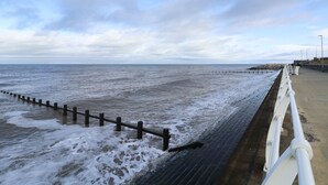A coastal walkway with a wooden barrier and a white railing, leading to the sea.