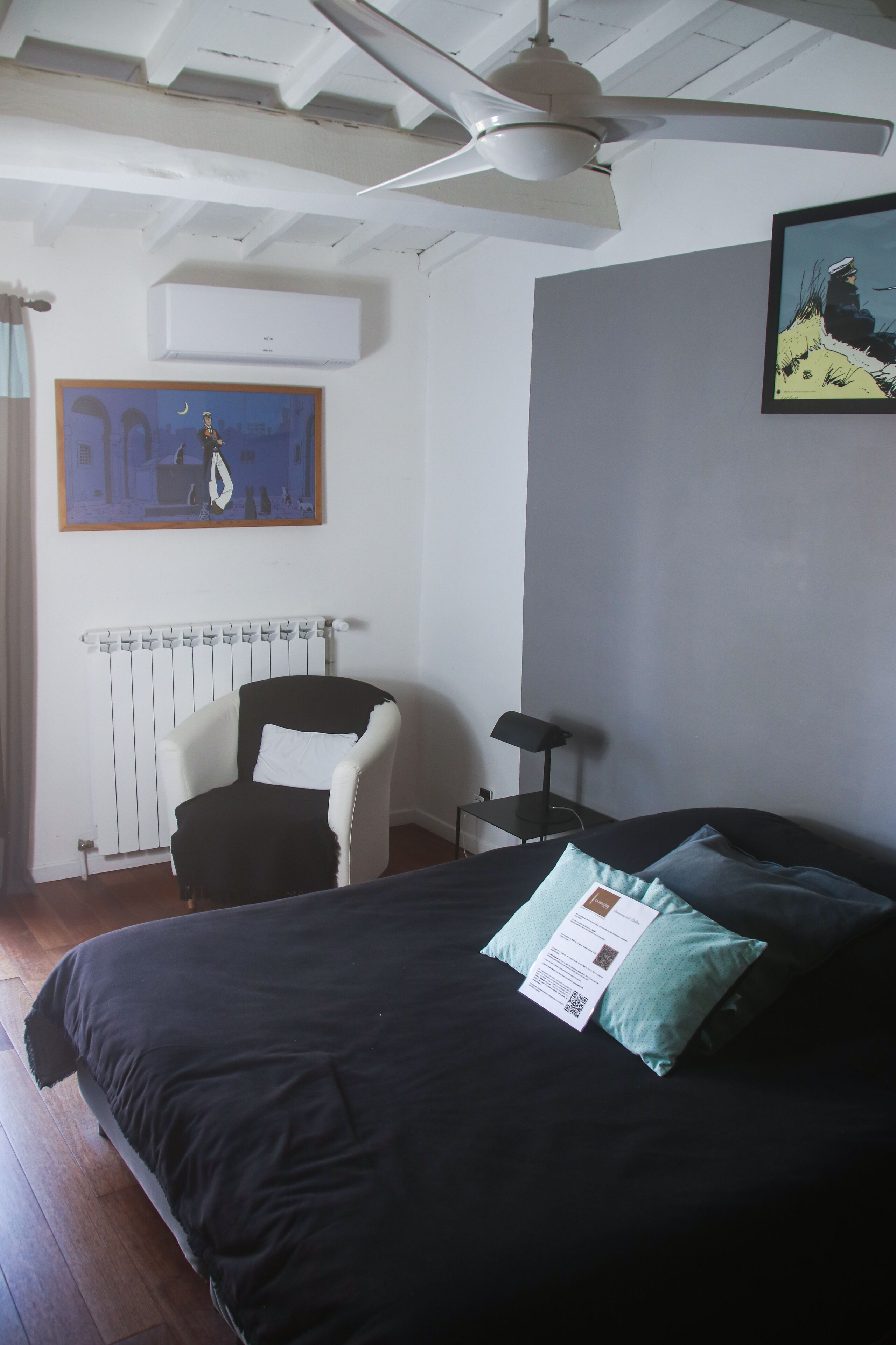 Une chambre à coucher avec un lit, une chaise, un radiateur, un ventilateur de plafond et un tableau accroché au mur.