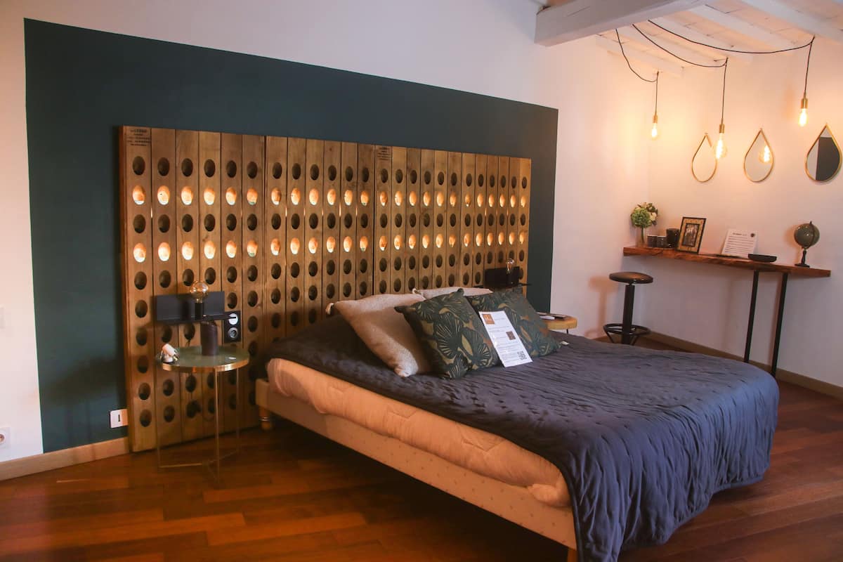 A bedroom with a wooden headboard, a bed with a dark blue bedspread, a small side table with a lamp, and a shelf with decorative items.