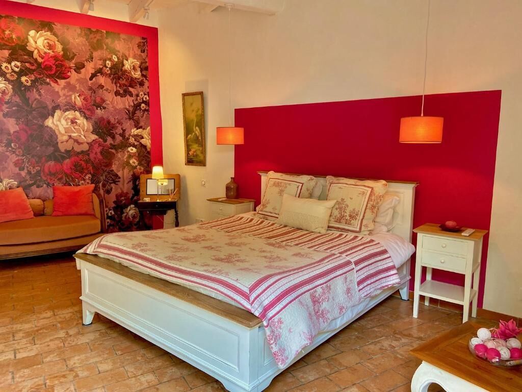 Un dormitori amb un llit gran, una paret decorada de color vermell, un tapet floral i una petita taula de nit de fusta amb una làmpada.