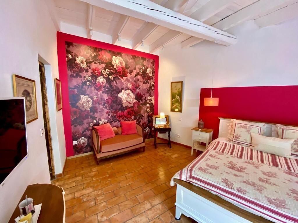 Un dormitorio con un papel tapiz floral grande, una cama con cubrecama a rayas, un sofá, una mesita con una lámpara y un televisor en la pared izquierda.