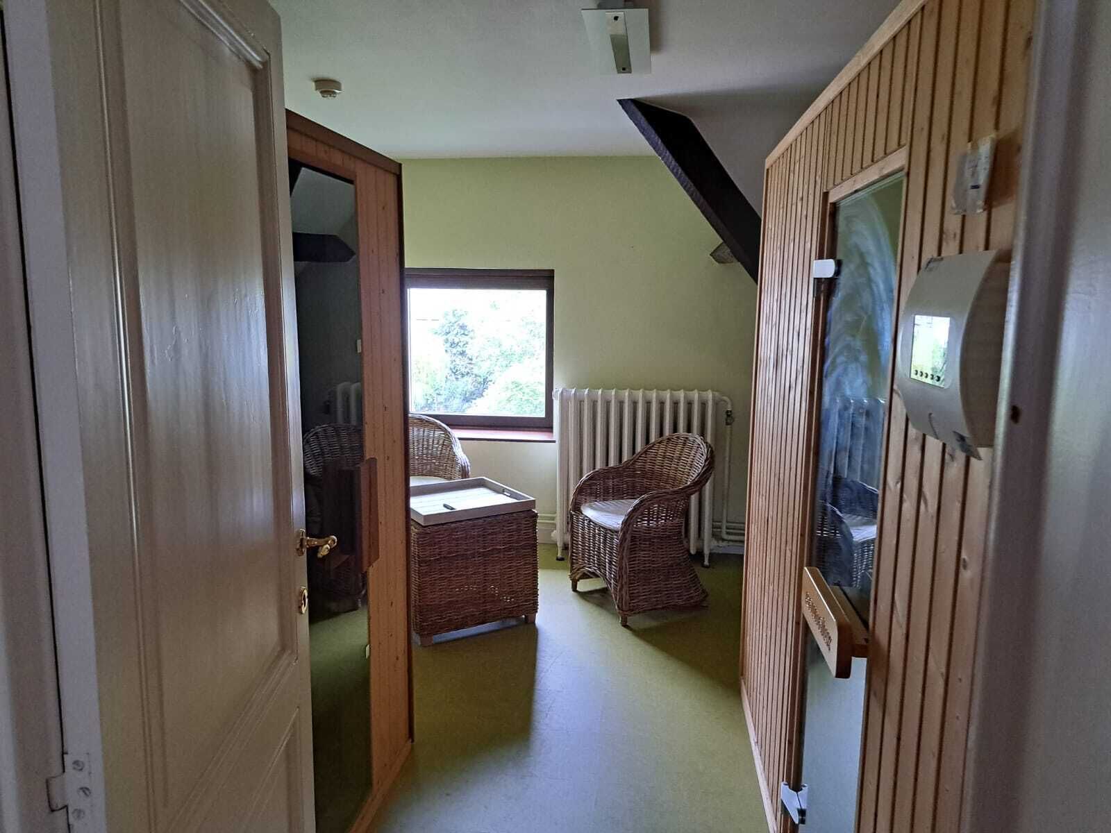 A narrow hallway with a wooden door, wicker chairs, a radiator, and a window with a view of trees.