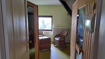 A narrow hallway with a wooden door, wicker chairs, a radiator, and a window with a view of trees.
