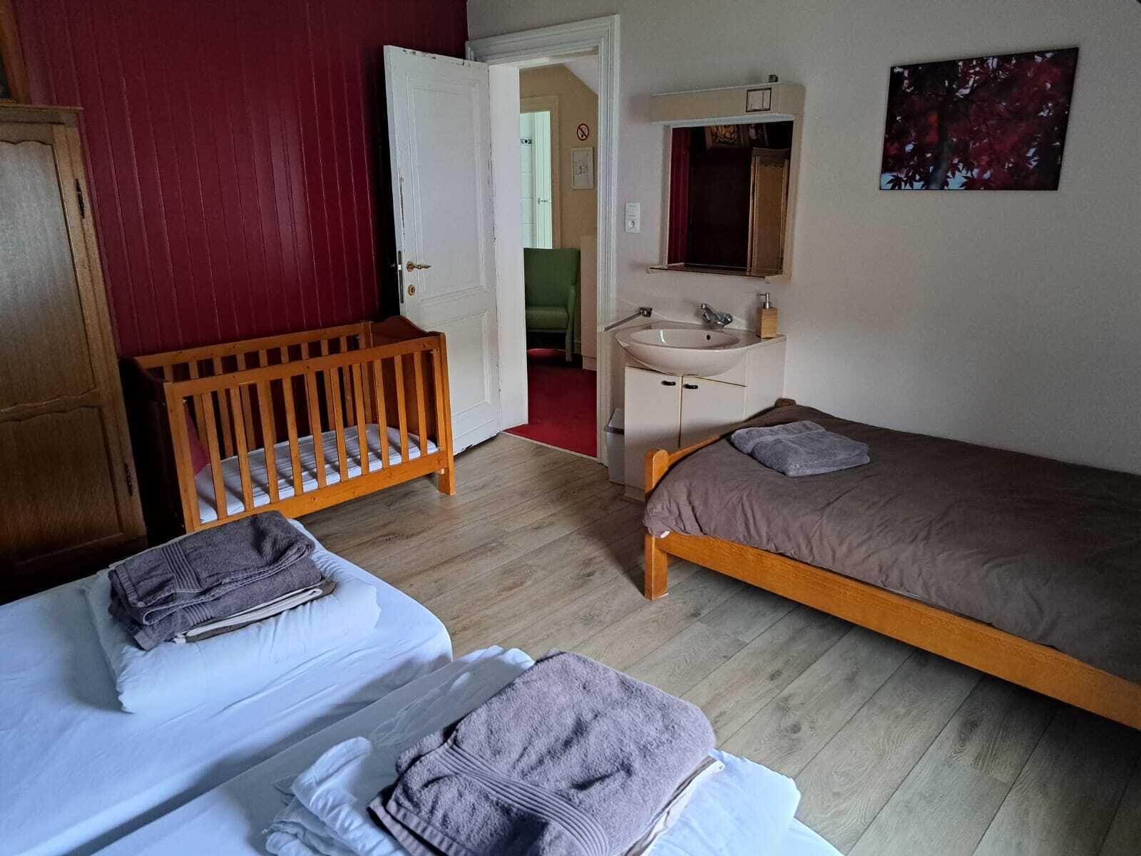 A bedroom with two beds, a wooden crib, a bathroom with a sink and mirror, and a red wall.