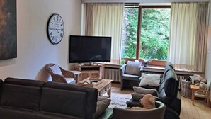 A living room with a sofa, a television, a clock, and a large window with curtains. - Villa Elie (Malmedy)